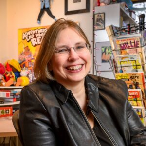 Smiling woman in a black leather jacket in a colorful comic-book shop behind her posters and shelves of comics visible